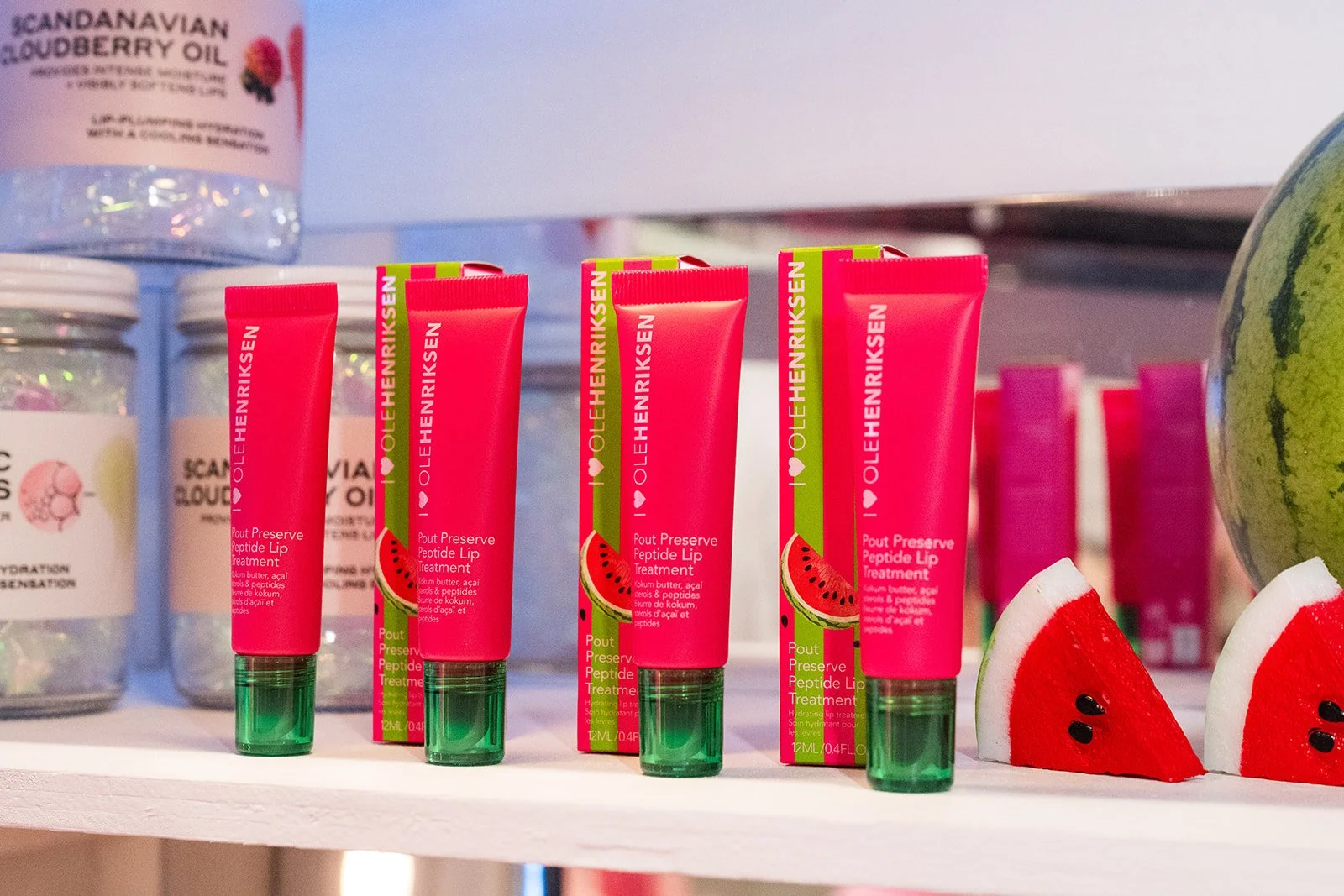 Several tubes of watermelon-flavored lip treatment with pink packaging and green caps, beside slices of watermelon on a store shelf.