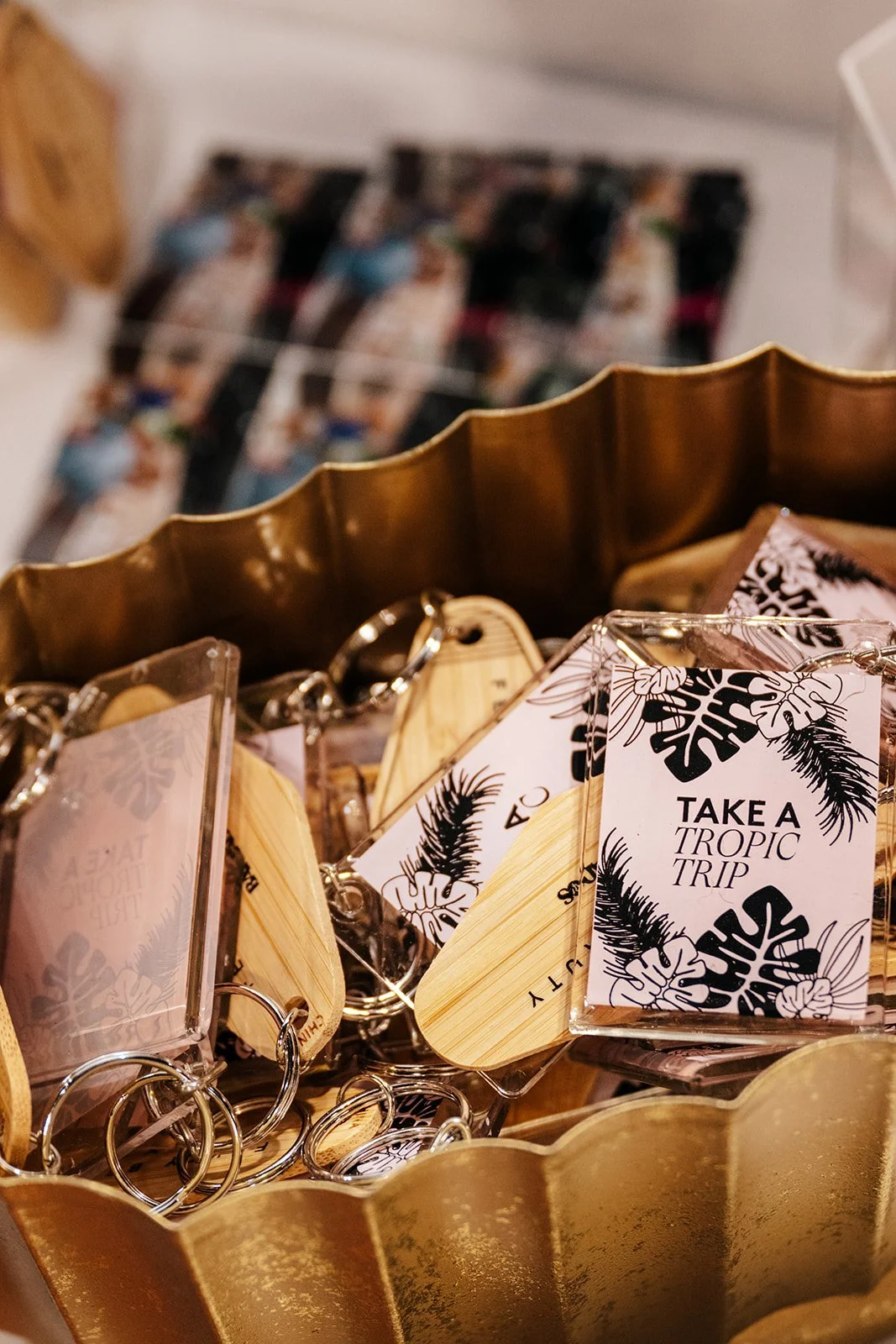 Close-up of keychains with tropical-themed tags reading "Take a Tropic Trip" in a decorative gold container, with a blurred photo of people in the background.