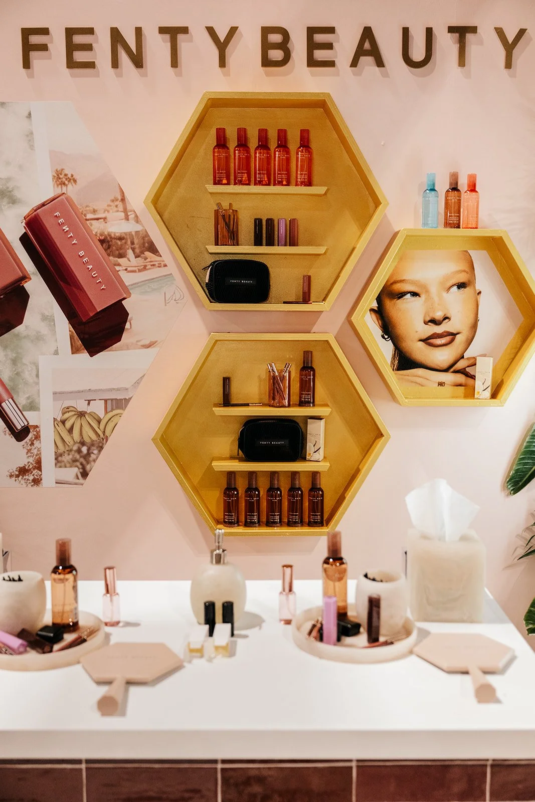 Display shelf for Fenty Beauty products with skincare bottles, makeup items, and promotional photos of a woman with smooth skin, set against a pink wall with the brand name at the top.