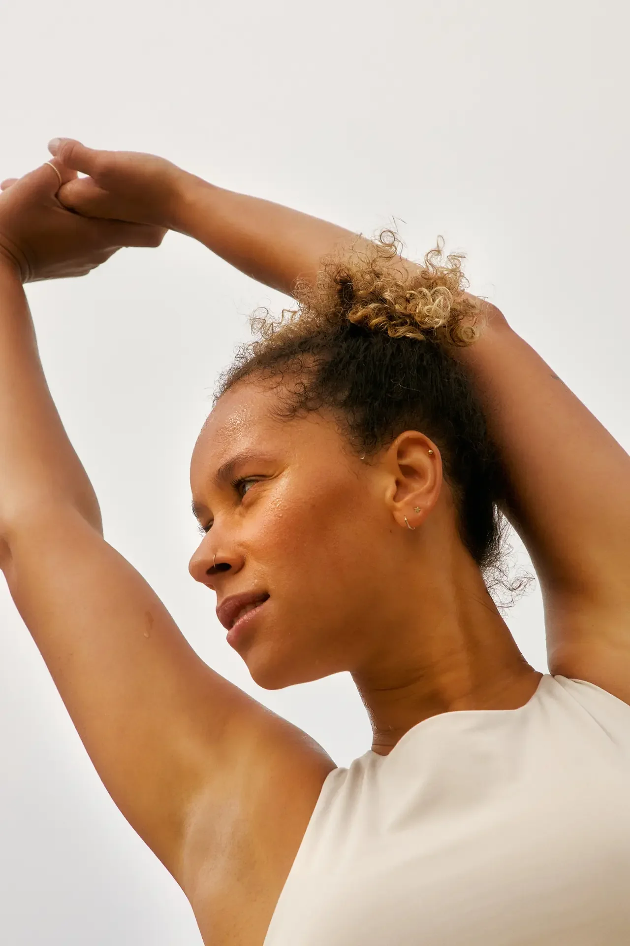 A woman with curly hair pulled up stretches both arms overhead against a plain light background, eyes lowered.