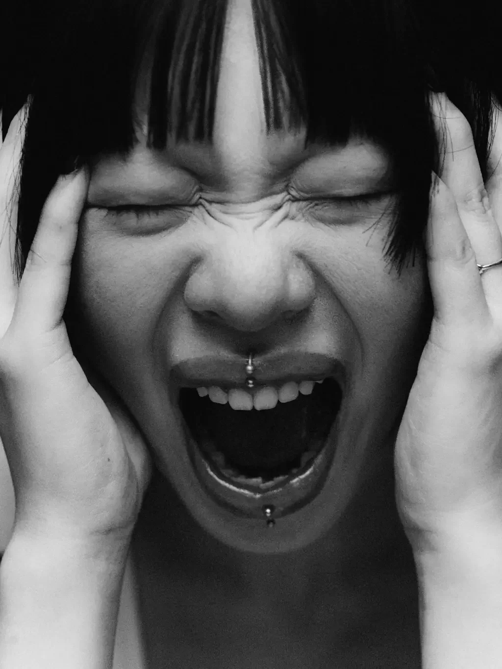 Black and white close-up of a woman with a fringe screaming with her eyes shut and her hands pressed to the sides of her head.