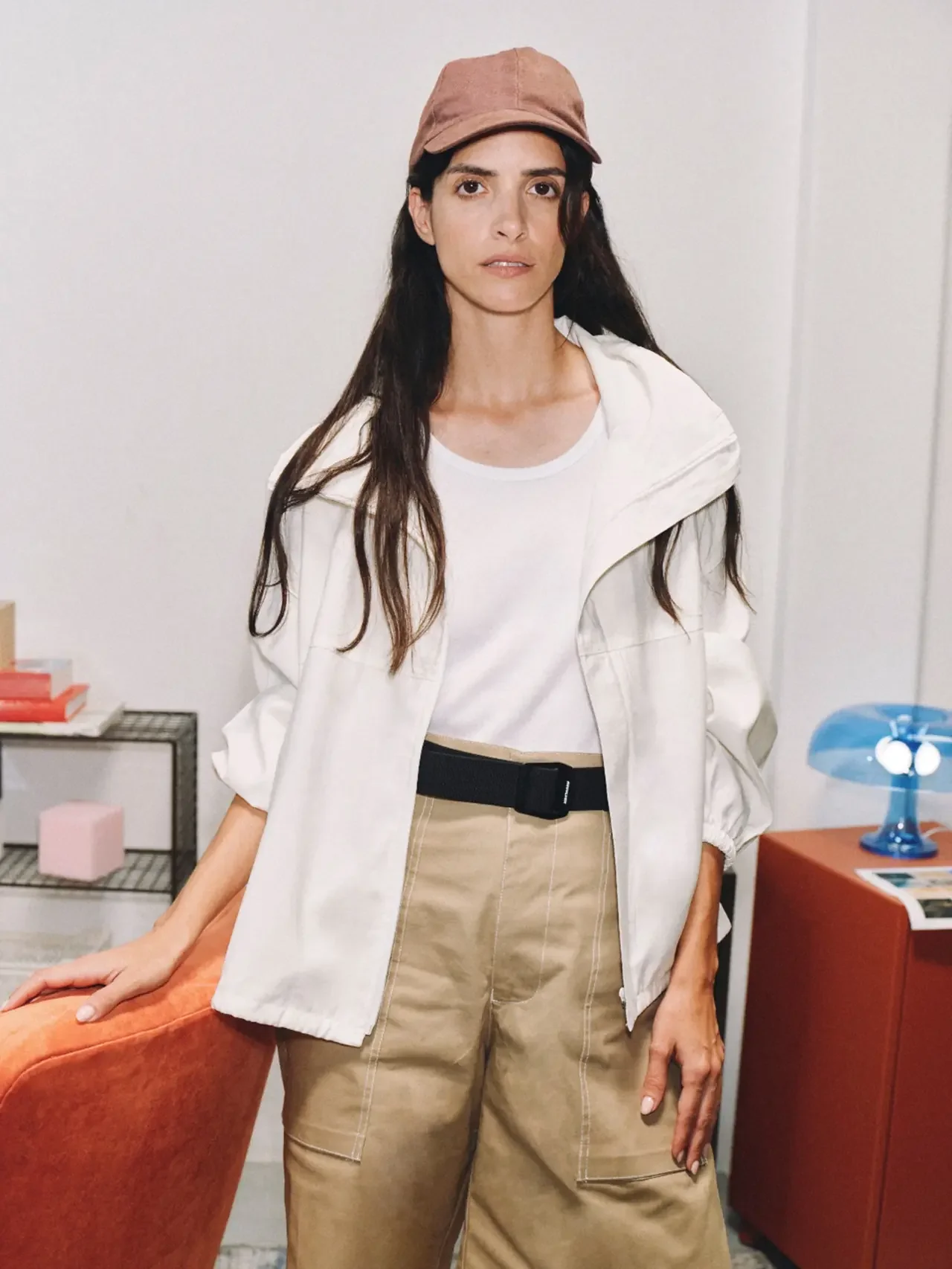 Woman in a brown cap, white jacket, and tan pants standing in a bright room beside an orange chair, looking directly at the camera.