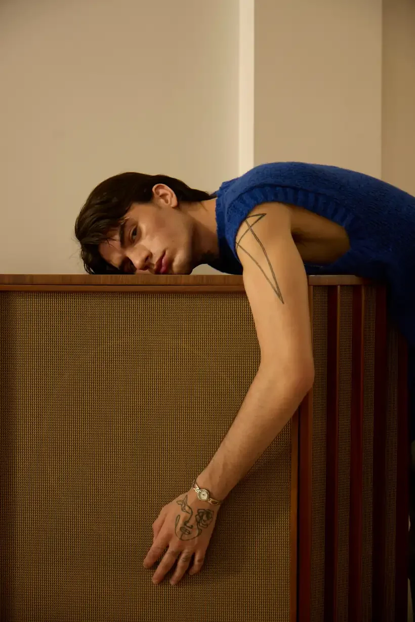 Exhausted young man in a blue knit vest draped over the top of a vintage wooden stereo cabinet, resting but not recharged