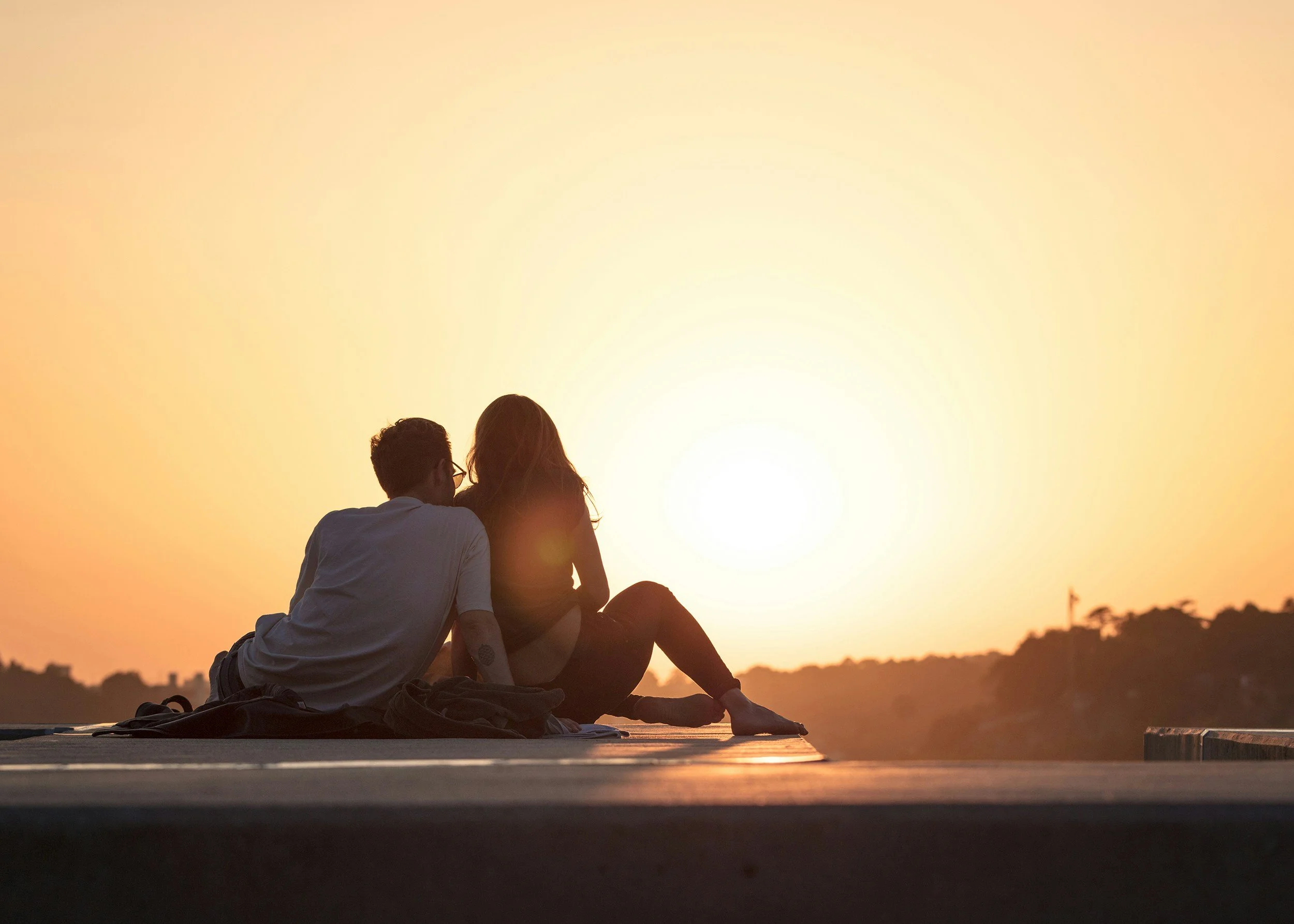 A couple sitting on a mat at sunset, with their backs to the camera, overlooking a scenic landscape.