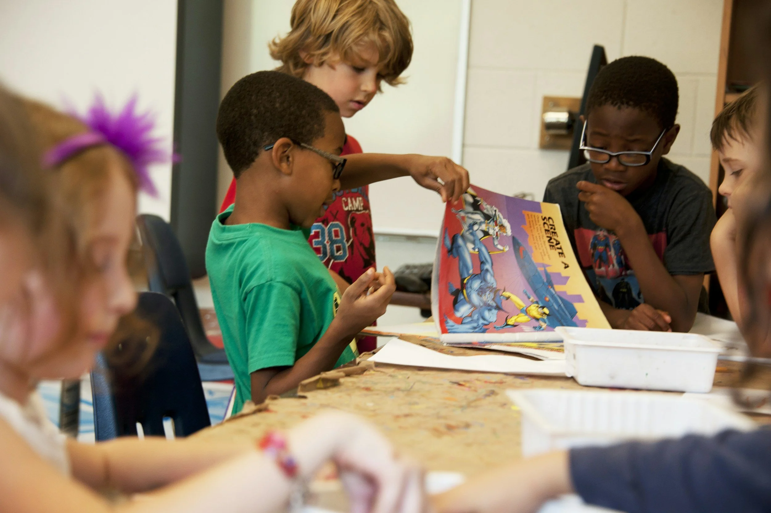 A group of children sitting around a table, with three of them looking at a comic book.