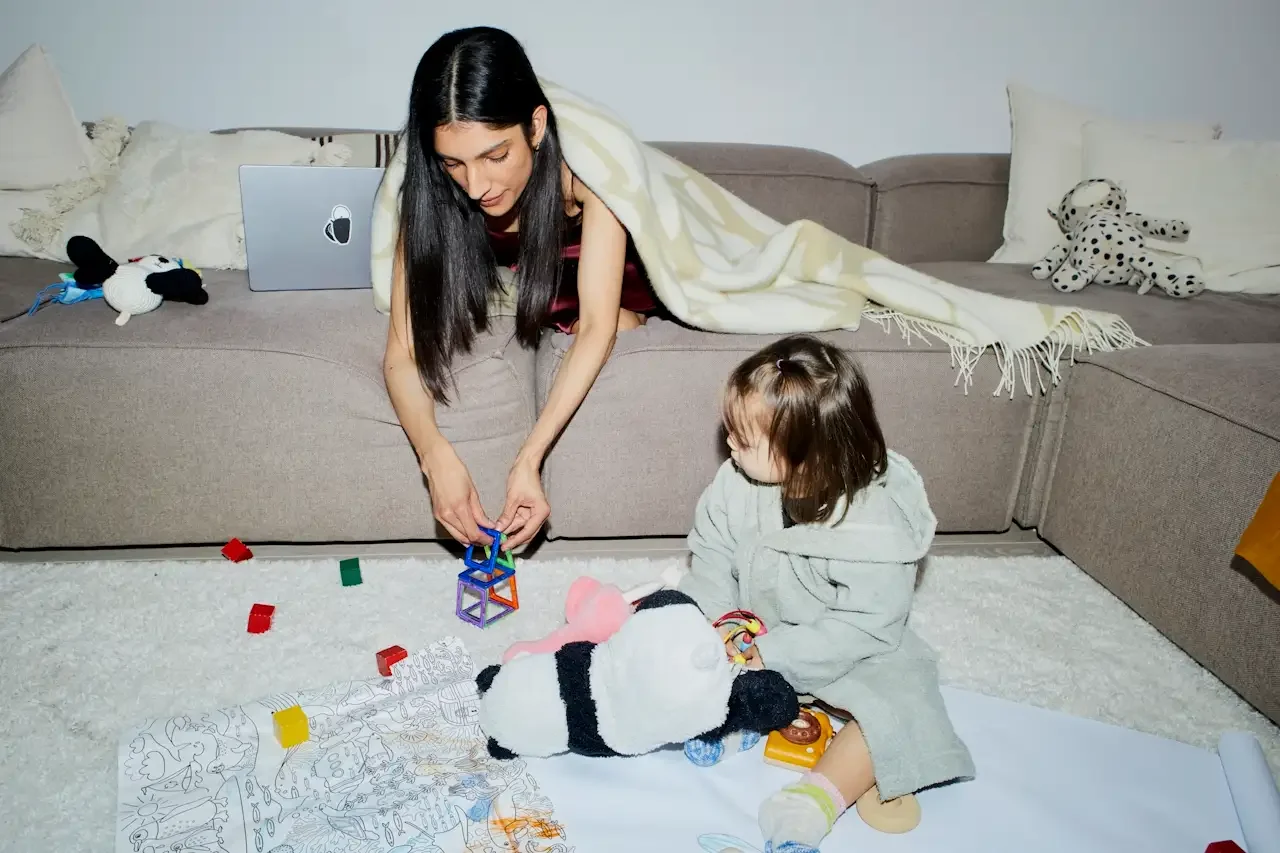 A mother working from home pauses to play magnetic tiles with her toddler on the living room floor, laptop open on the couch behind her, illustrating the constant context-switching of parenting with ADHD during perimenopause.