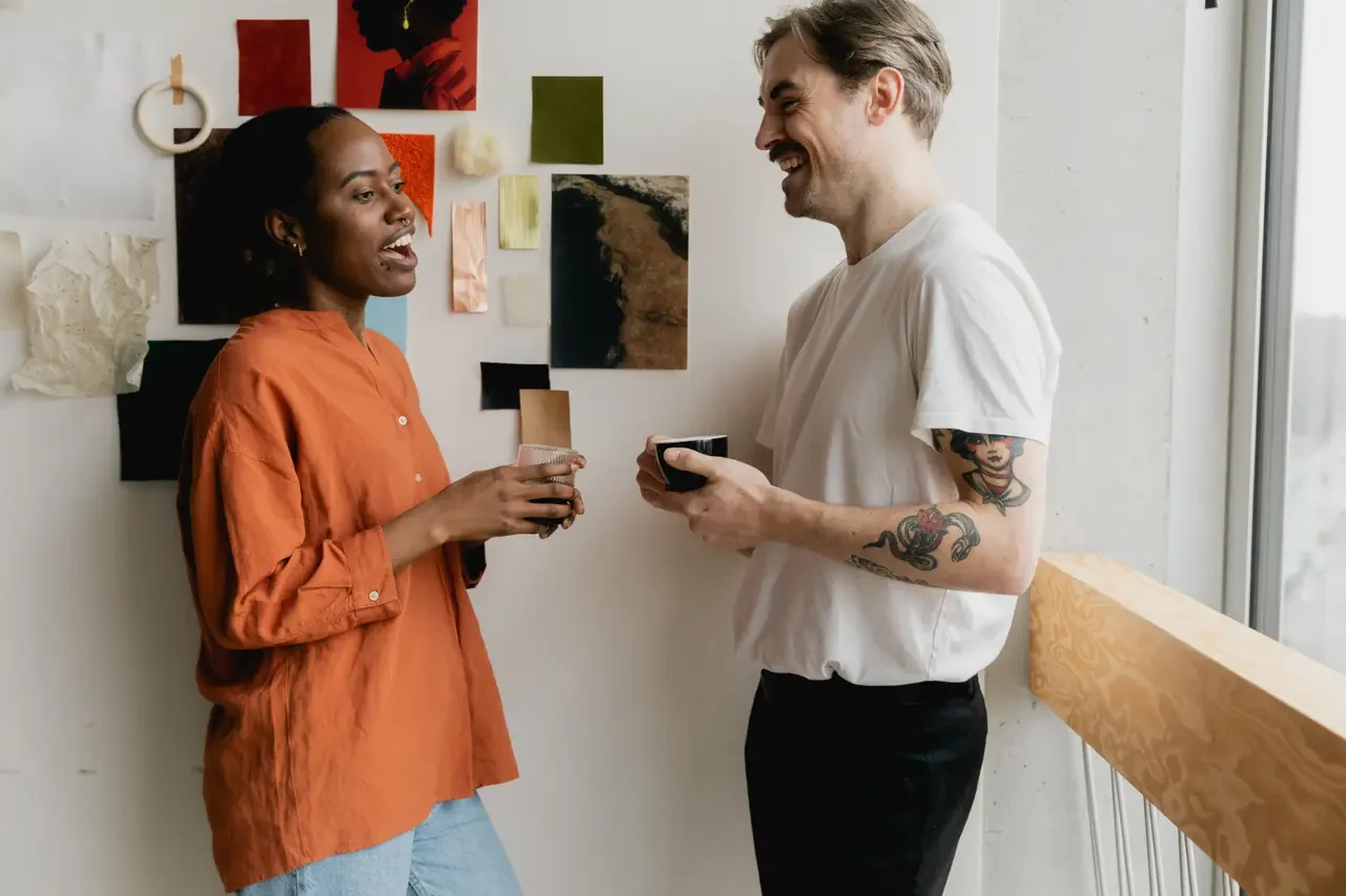 A woman in an orange button-down shirt and a man in a white t-shirt stand facing each other holding cups, laughing, beside a wall covered in fabric and paper swatches.