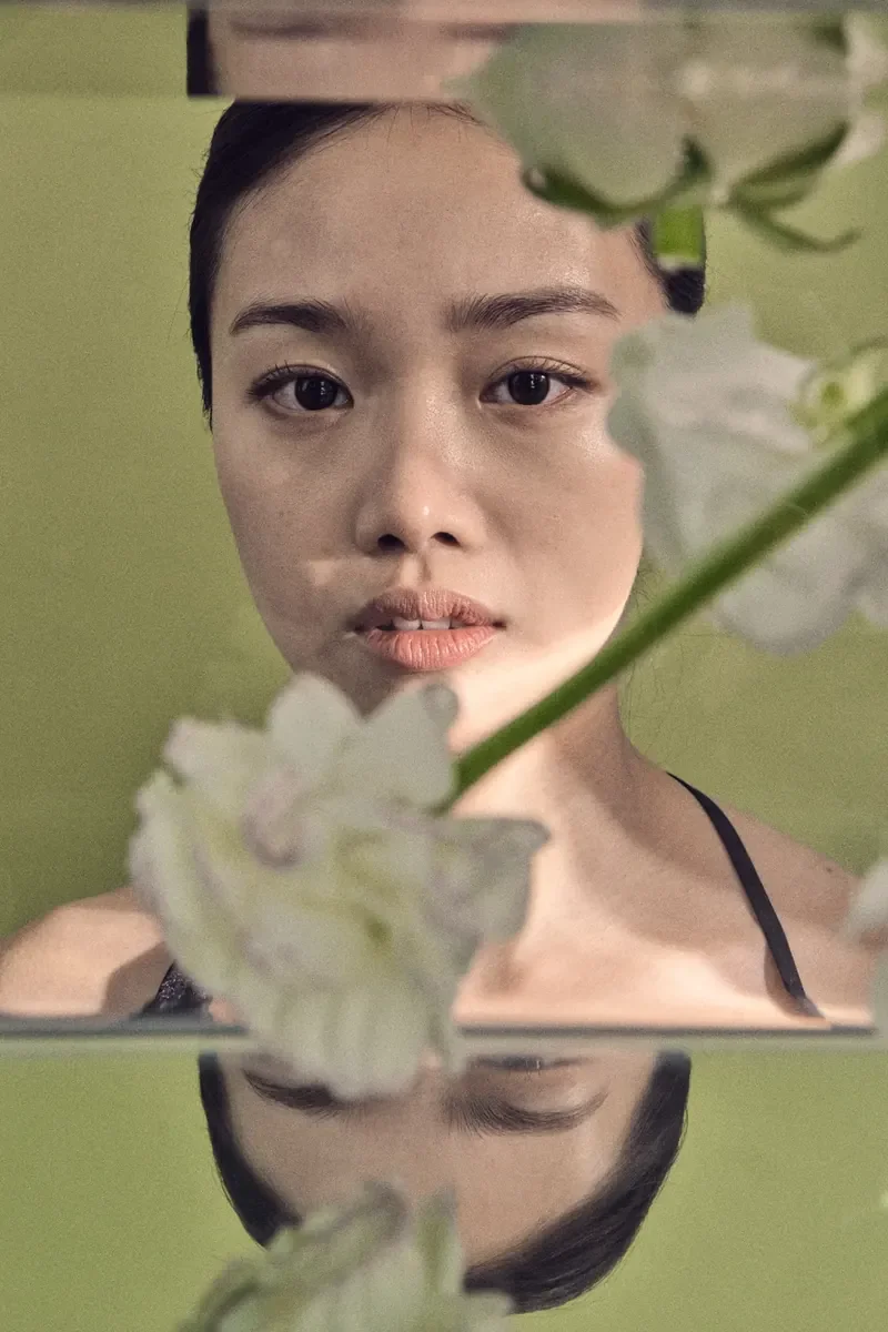 Woman looking into a mirror behind blurred white flowers, with her face reflected upside down in a second mirror below.
