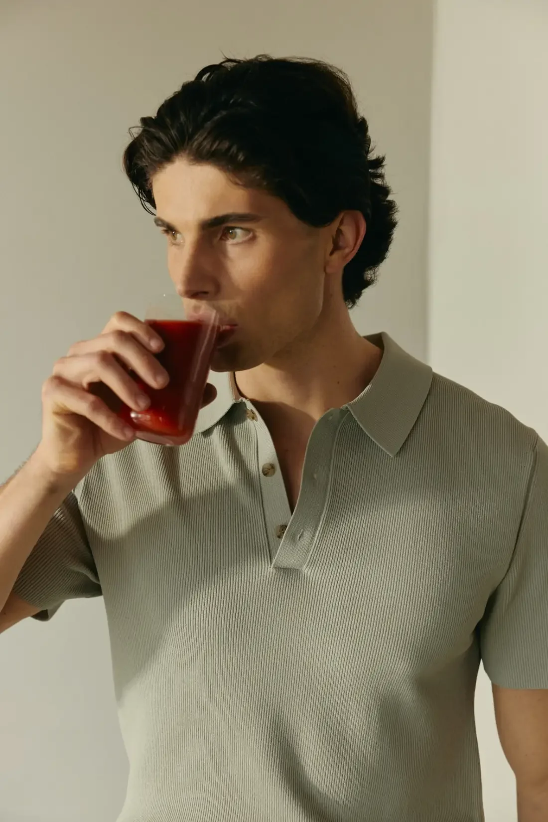 Man in a sage green knit polo sipping a red drink and gazing off to the side, a quiet pause in anxiety and ADHD care