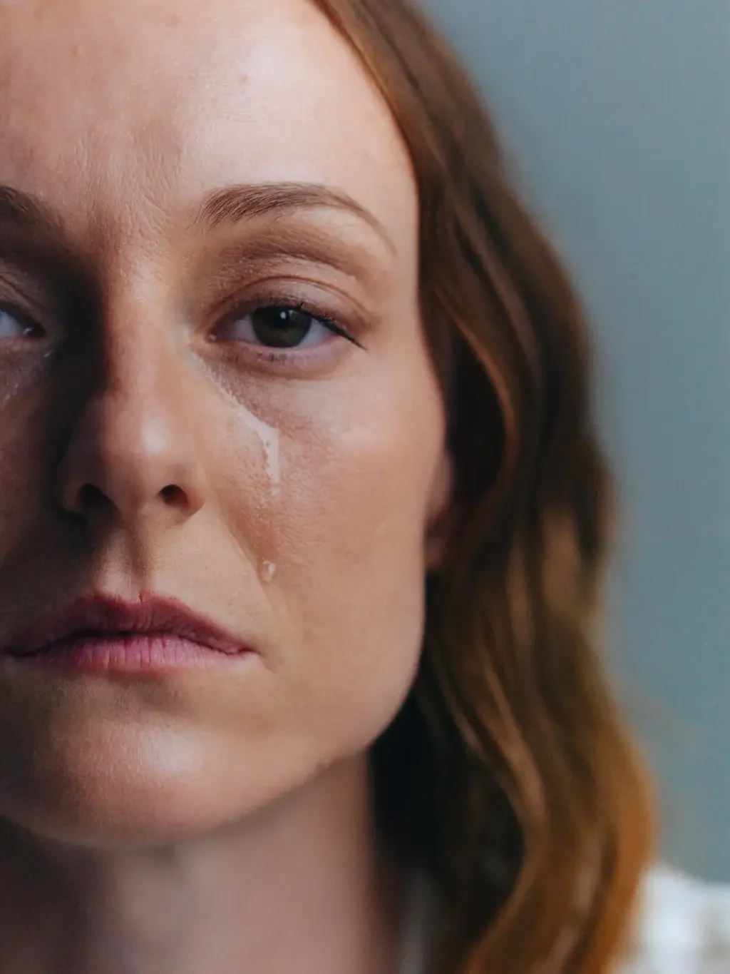 Close-up of a woman's face with a tear running down her cheek, looking away from the camera against a grey-blue wall.