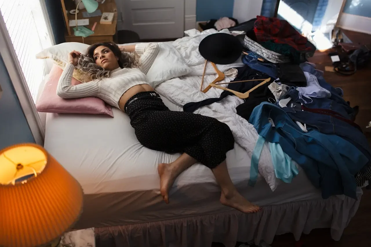 Adult woman lying on an unmade bed surrounded by piles of unfolded laundry and empty hangers, illustrating the awareness-without-execution pattern in adults with ADHD perfectionism.