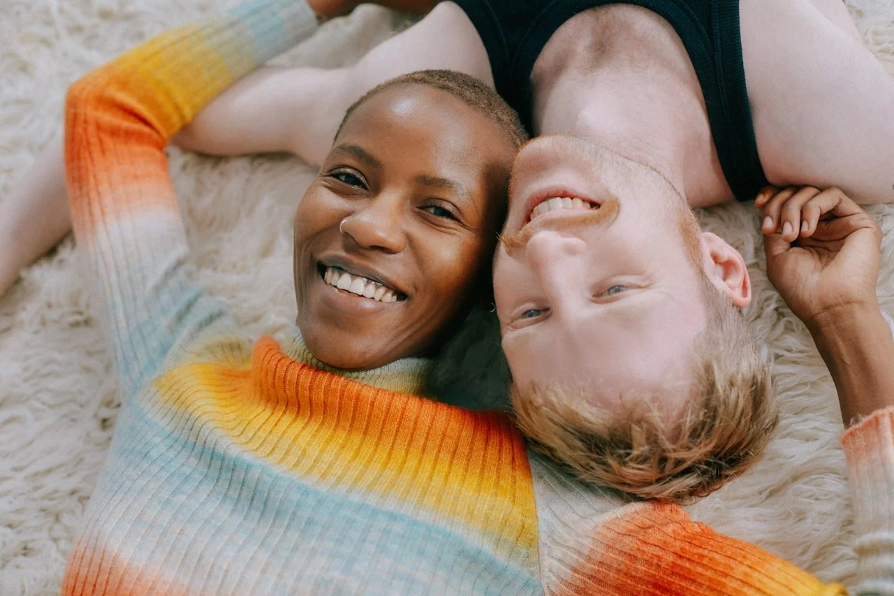 Two people, a woman with dark skin and a man with light skin, lying on a fluffy white rug, smiling and looking at the camera.