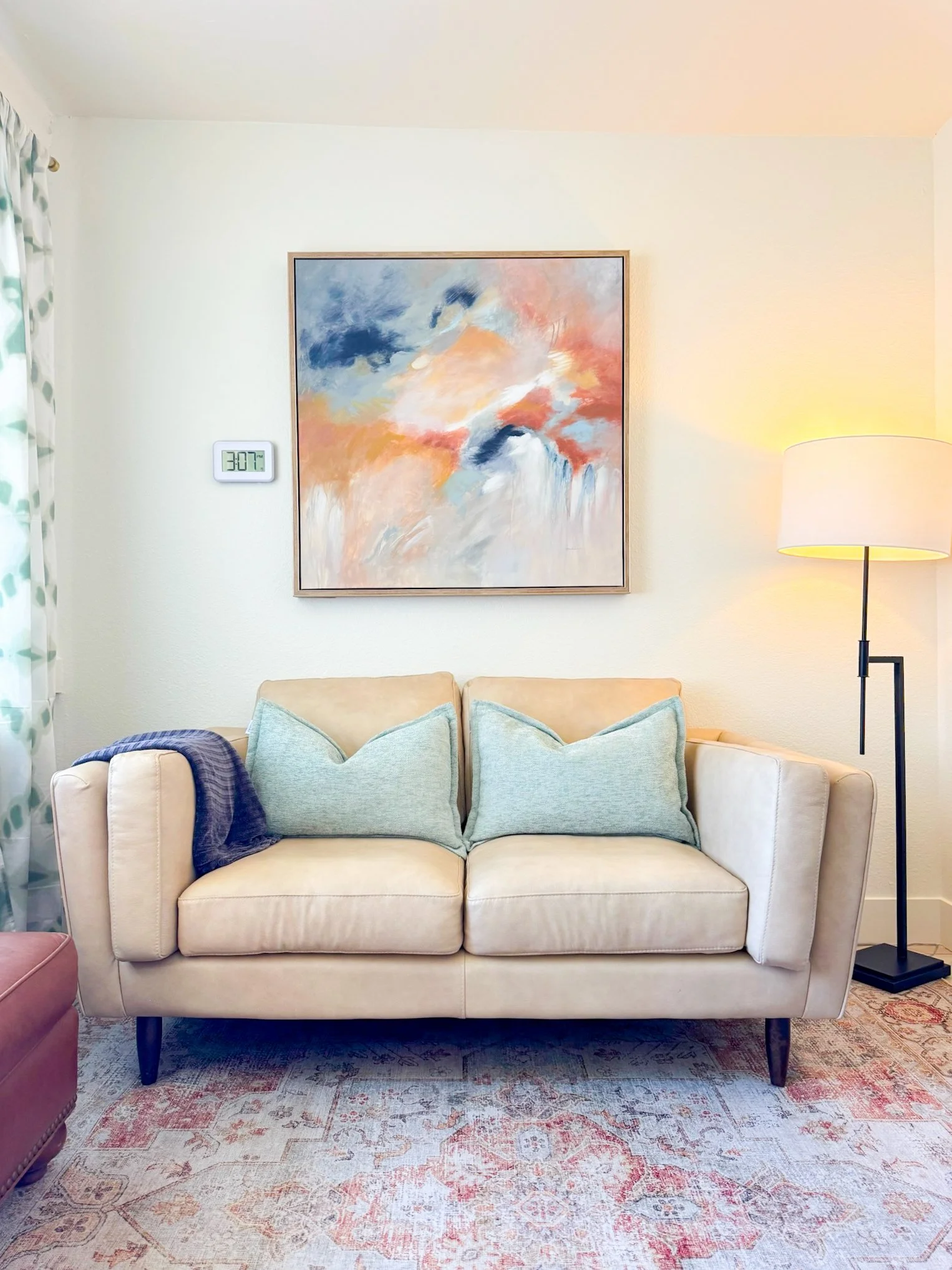 A cozy therapy room corner featuring a beige sofa with mint green cushions, a dark blue throw blanket on the arm, a colorful abstract painting on the wall, a modern tall floor lamp, and a patterned rug on the floor.