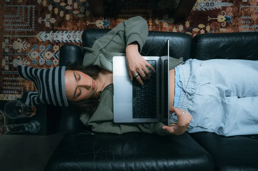 Overhead view of a woman lying on a black leather couch with her eyes closed and an open laptop resting on her torso, one hand still on the keyboard.