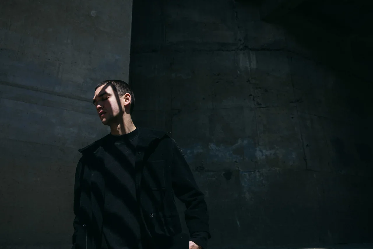 A man in a black jacket stands against a concrete wall, eyes closed, with harsh diagonal shadows falling across his face.
