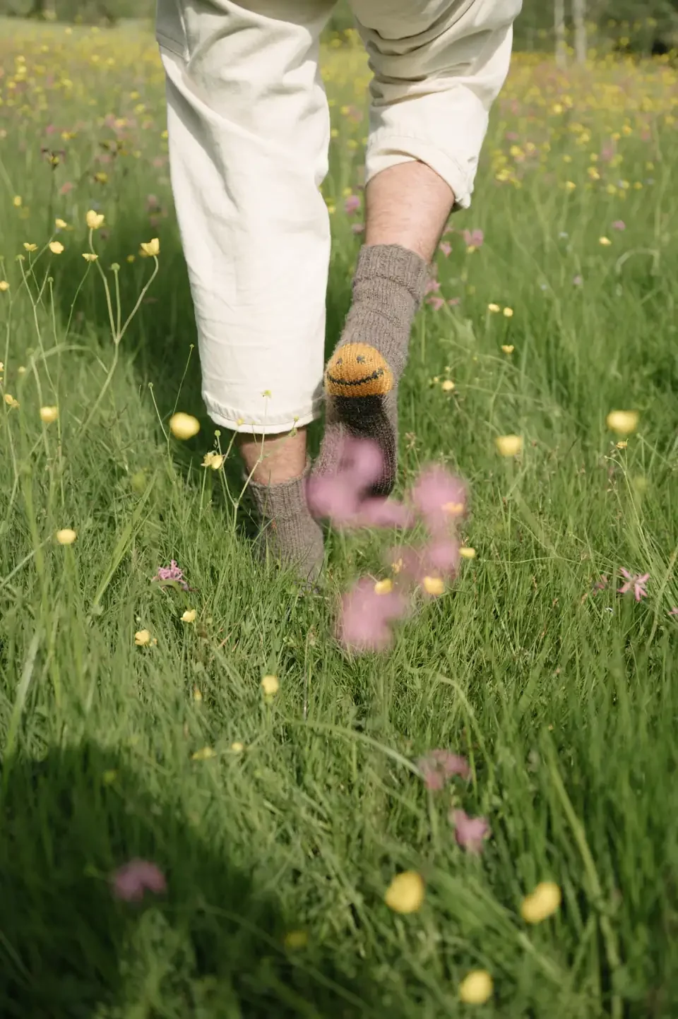 A person in cream trousers and gray wool socks with a yellow smiley face on the heel, walking through a meadow of yellow and pink wildflowers.
