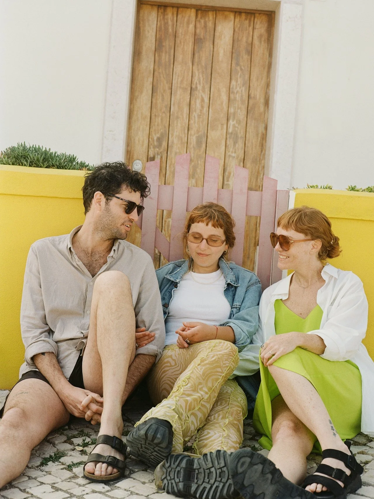 Three people sitting on cobblestone ground by a yellow wall, with a pink fence and wooden door in the background, enjoying each other's company.