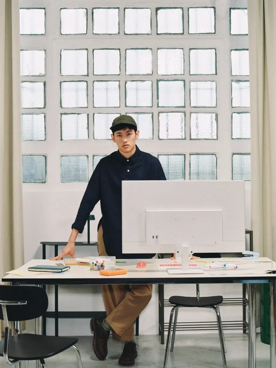 Person in a cap and dark shirt standing behind a white desk with a monitor, colorful markers, and a notebook, with a glass-block window behind them.