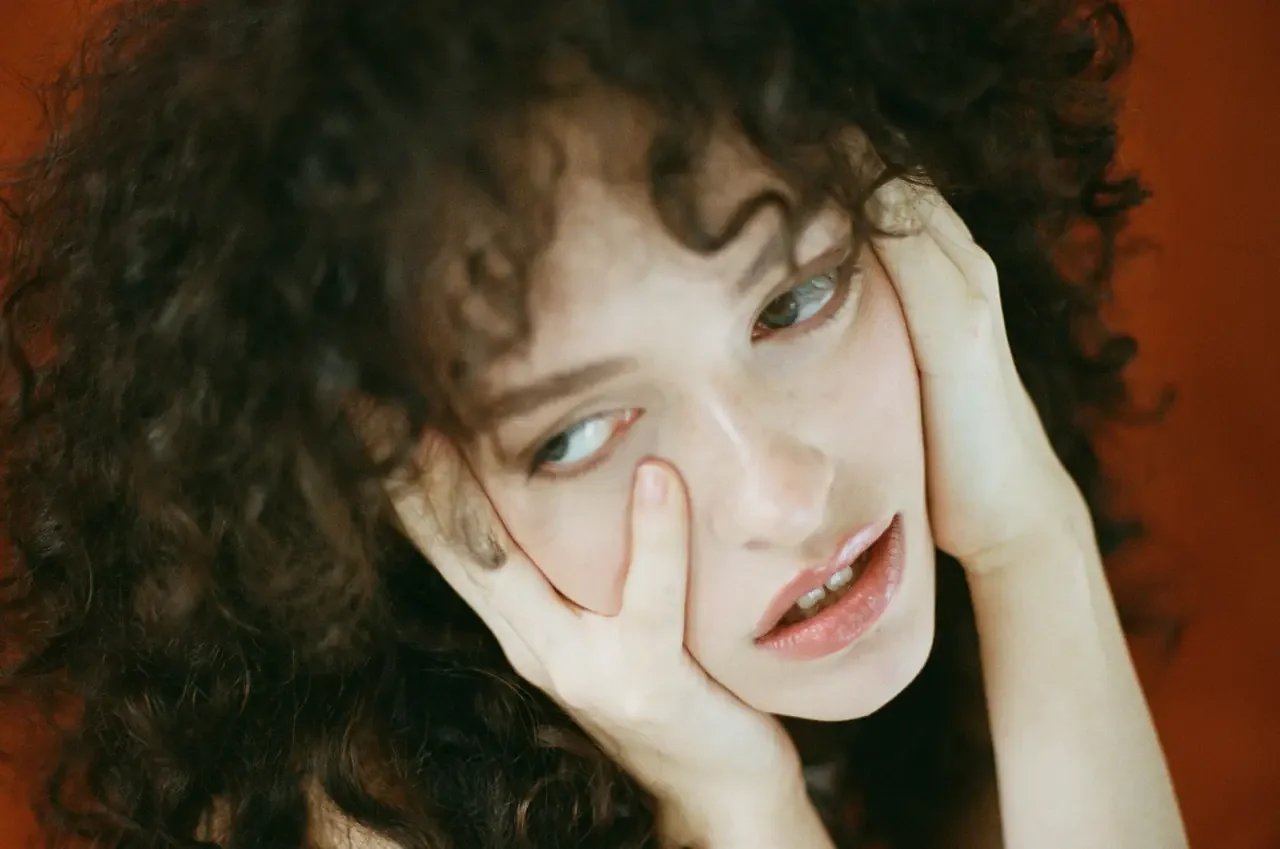 A woman with dark curly hair rests her cheek against her hand, looking away, against a warm orange background.