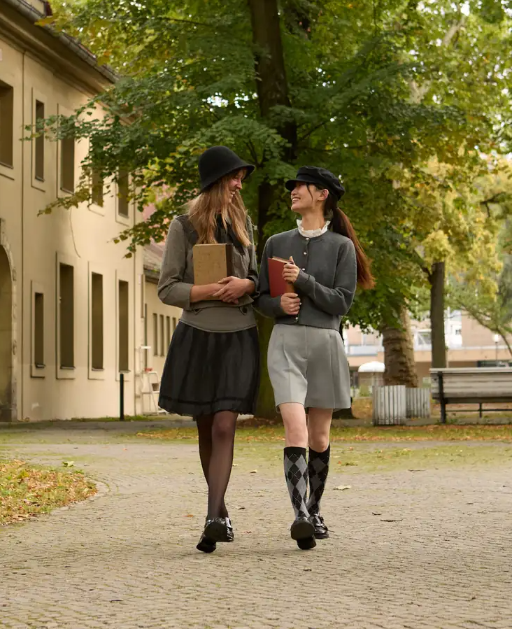 Two women in hats carry books as they walk side by side along a cobblestone path lined with trees.