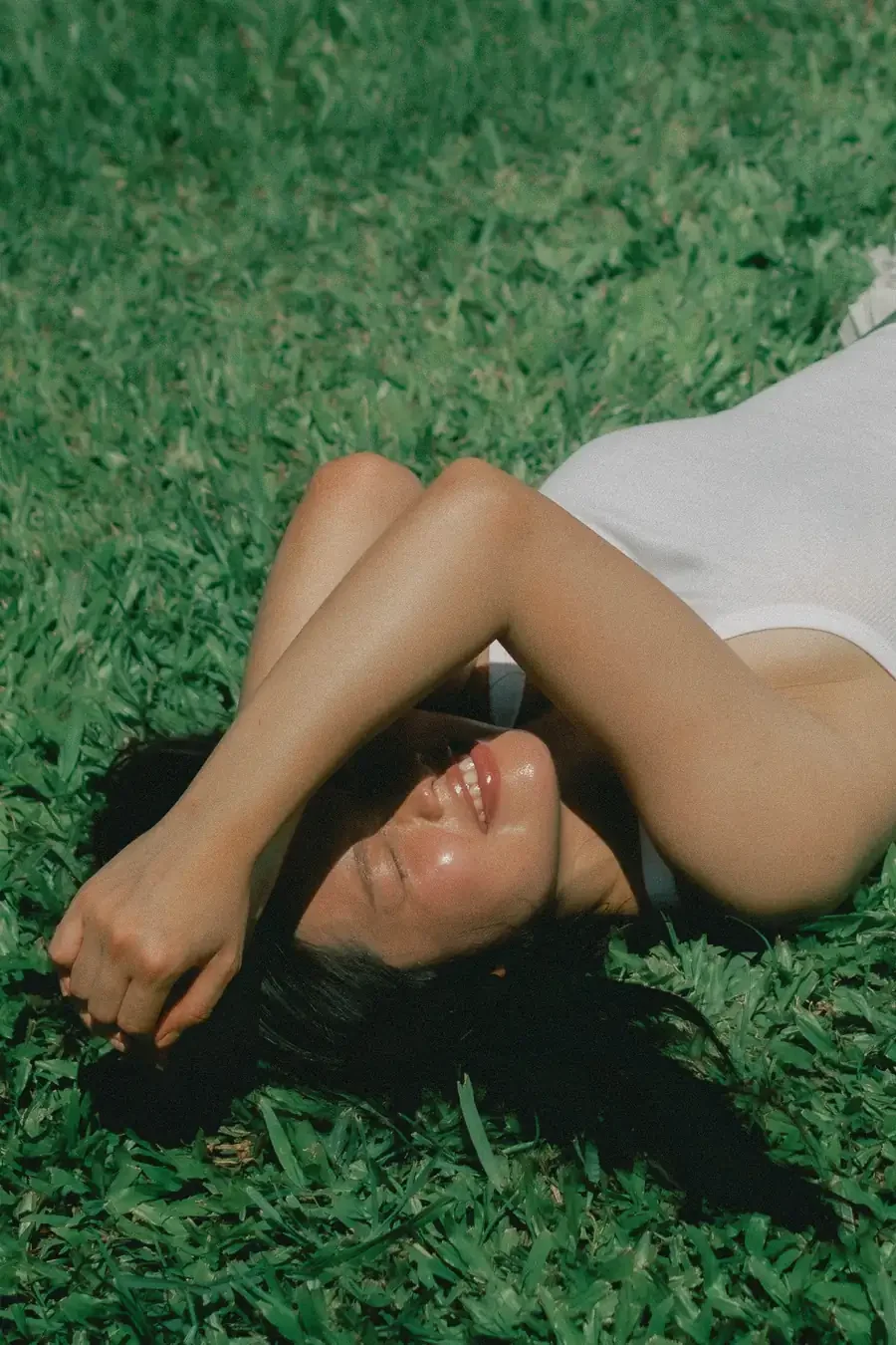 A woman lies on her back in sunlit grass with one arm resting across her eyes, smiling.