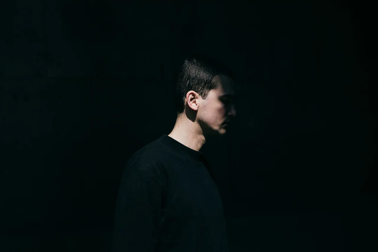 Side profile of a man in a black sweater against a dark background, light falling across half his face as he looks down.