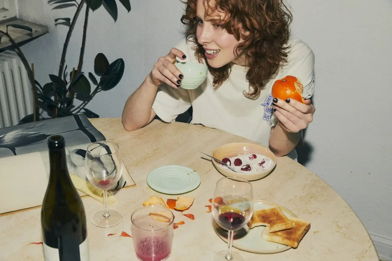 Smiling woman with curly red hair holds a teacup and a half-peeled orange at a kitchen table scattered with dishes, glasses, toast, and orange peels.