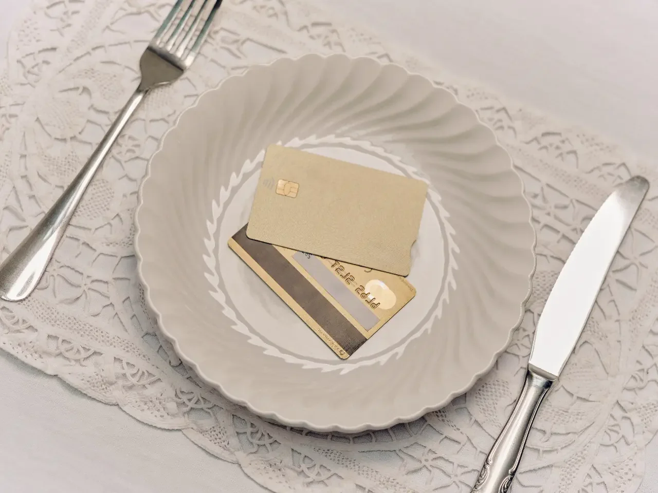 Two bank cards resting on a fluted white dinner plate at a formal place setting with a fork, knife, and lace placemat.