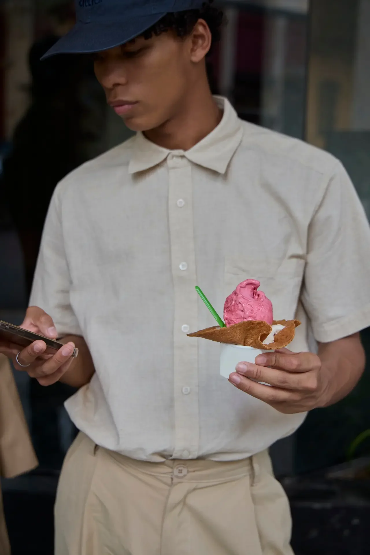 A person in a linen shirt and cap holds a cup of gelato in one hand while looking down at a phone in the other.