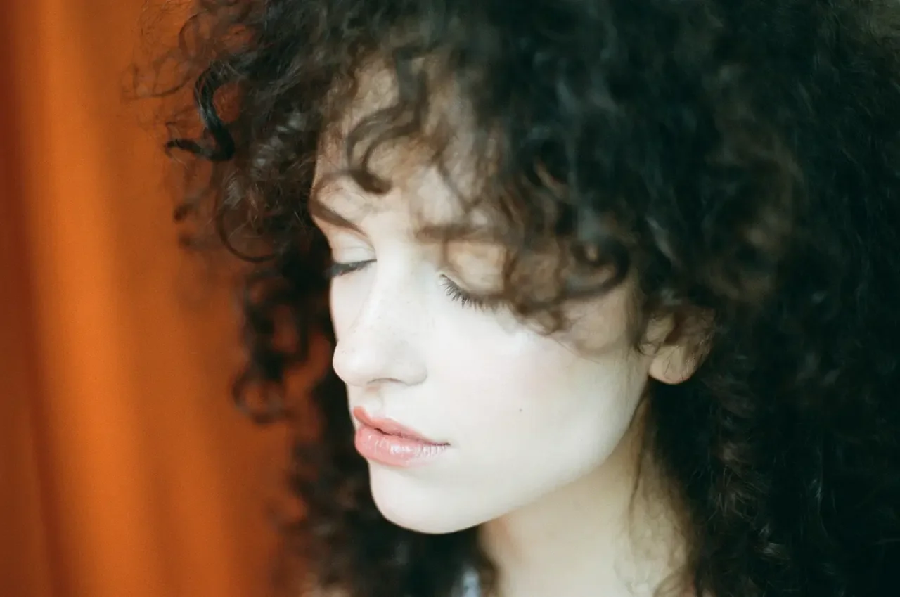 Side profile of a woman with dark curly hair and freckles, eyes closed, beside an orange curtain.