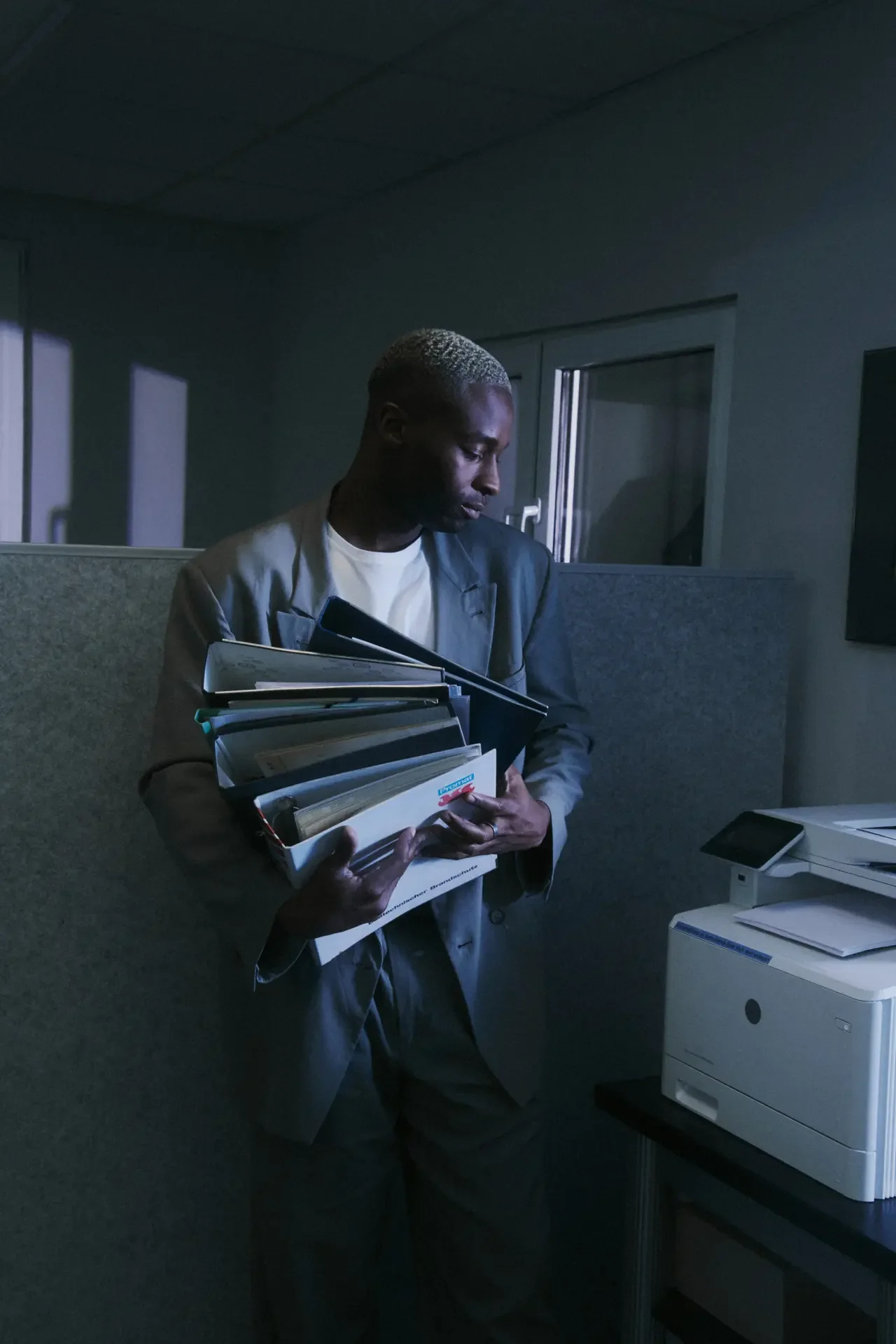 Man in a suit carrying an overloaded armful of binders and folders through a dim office, weighed down by an invisible workload
