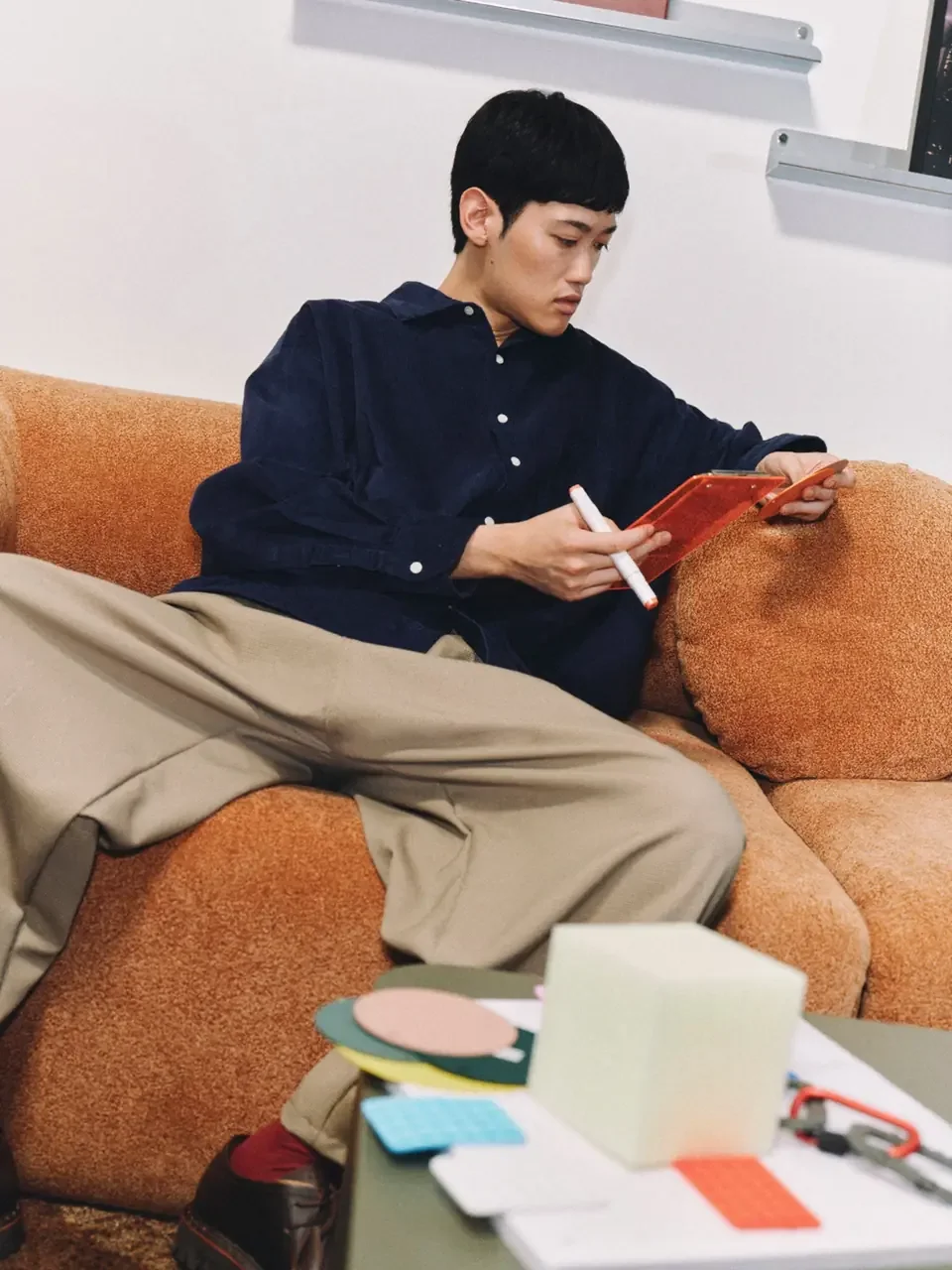 Person in a navy corduroy shirt sitting on an orange couch, holding a marker and reading notes on a clear orange clipboard.