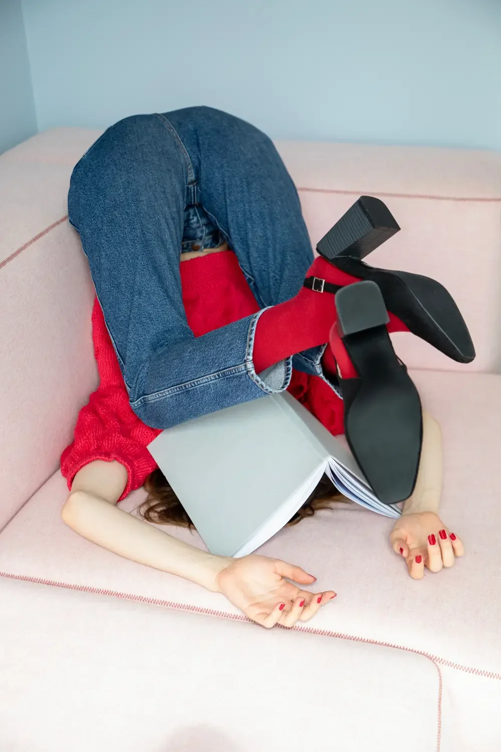 Woman in a red sweater and jeans lying upside down on a pink couch with an open book covering her face, a common image of ADHD overwhelm in women