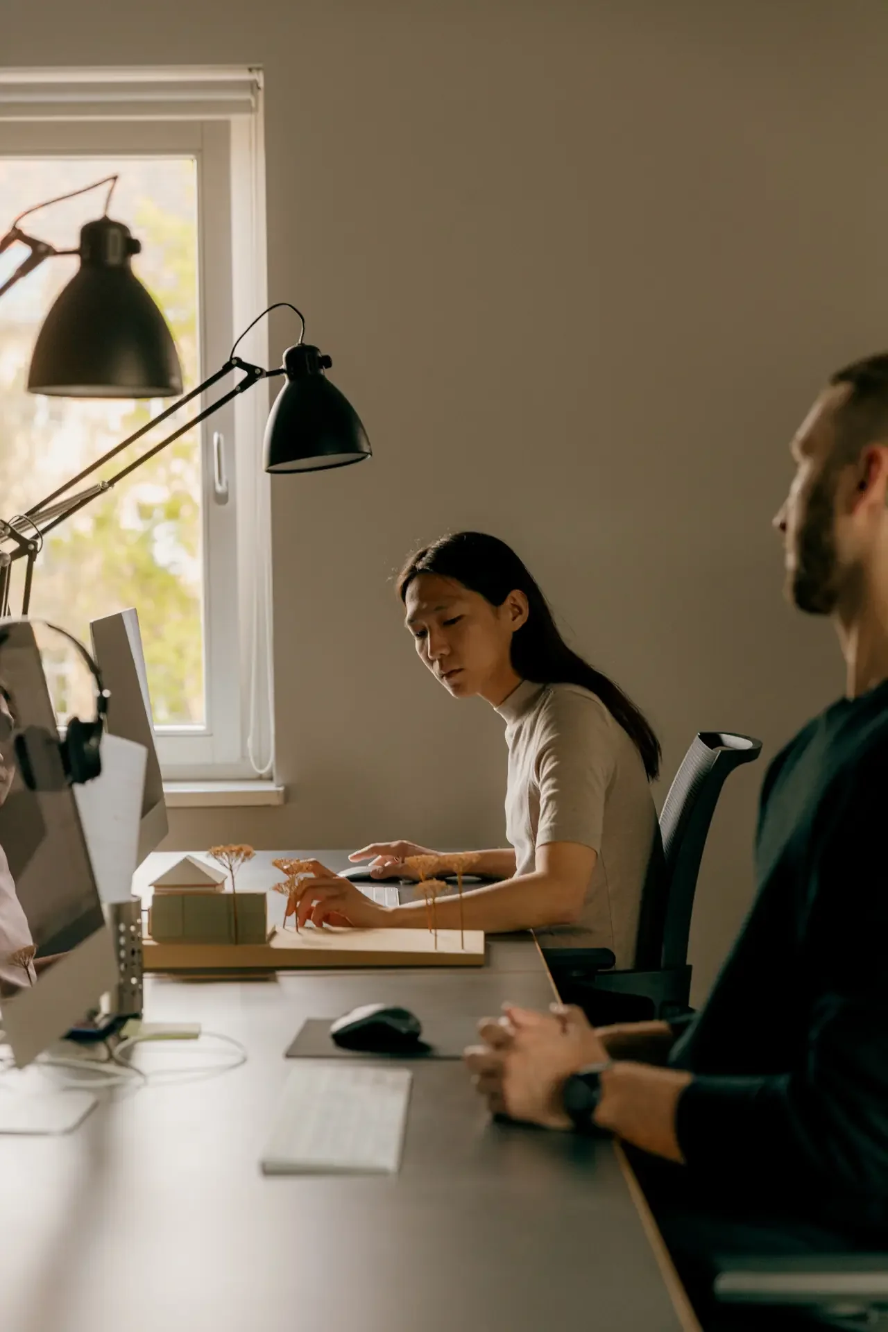 Person working intently at a shared studio desk in low evening light, coworker in the foreground — the quiet overwork behind high-functioning ADHD