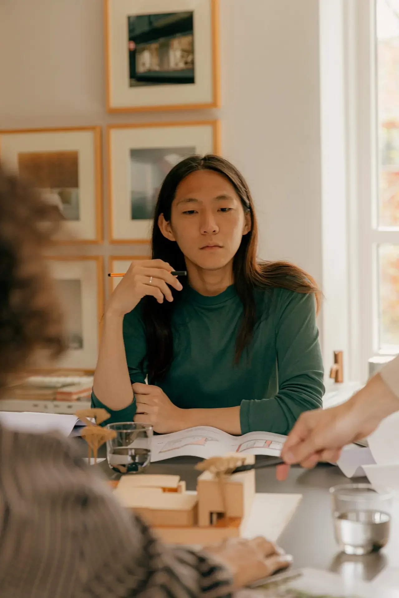Person in a green sweater sitting attentively in a meeting, pen in hand, colleagues gesturing at plans across the table — the composed performance of high-functioning ADHD