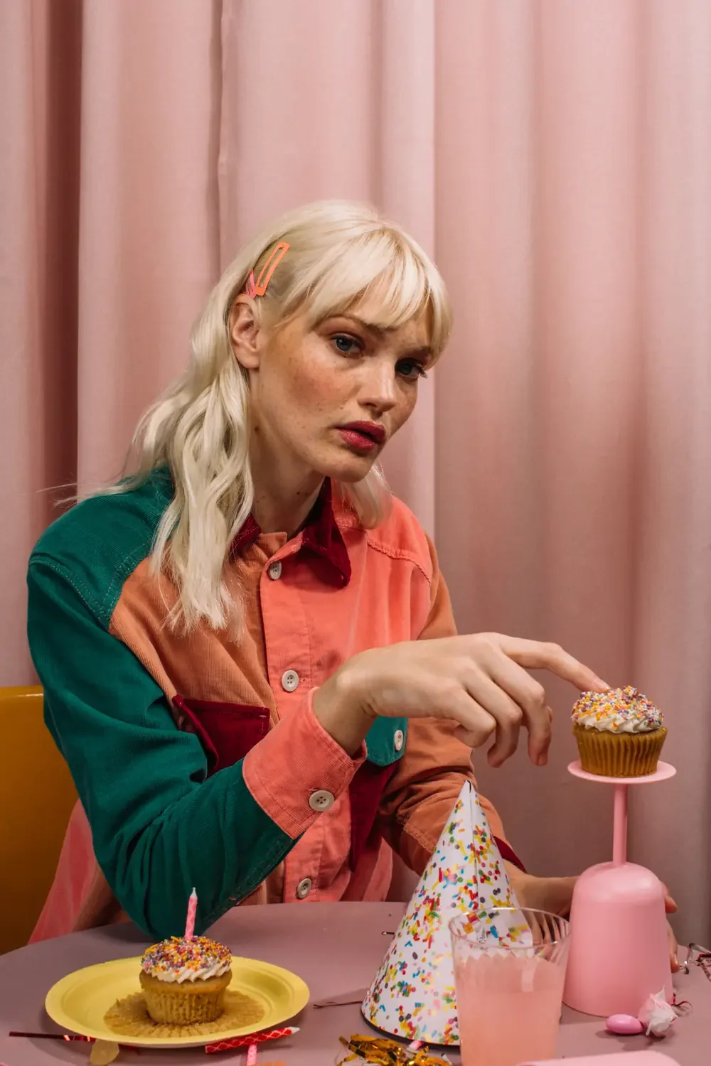 Woman with blond hair sitting alone at a pink birthday table, pressing one finger into a cupcake, showing rejection sensitive dysphoria in women with ADHD