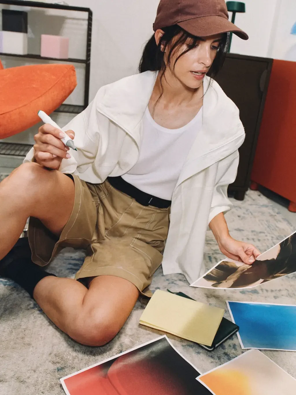 Woman in a brown cap and white jacket sitting on a rug, holding a marker and looking at a photo print, with more colorful prints and a notebook spread on the floor around her.