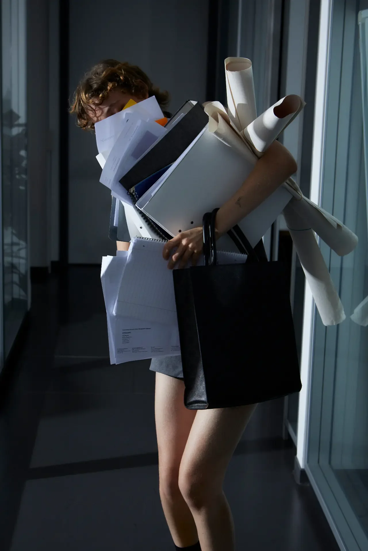 Woman in an office hallway struggling to hold binders, loose papers, rolled posters, and a tote bag, showing how ADHD overwhelm builds at work
