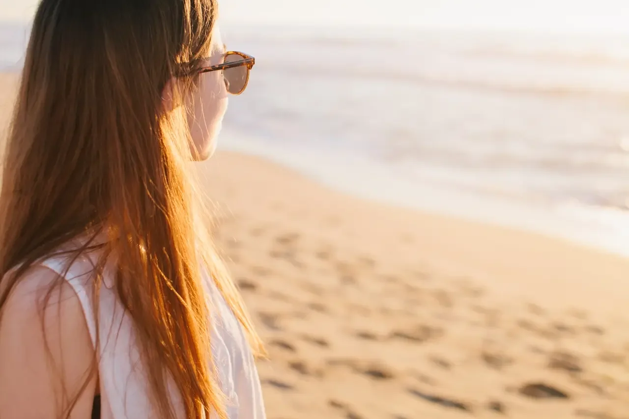 Woman with long hair and sunglasses looking out at the ocean at golden hour, reflecting quietly on a beach in California