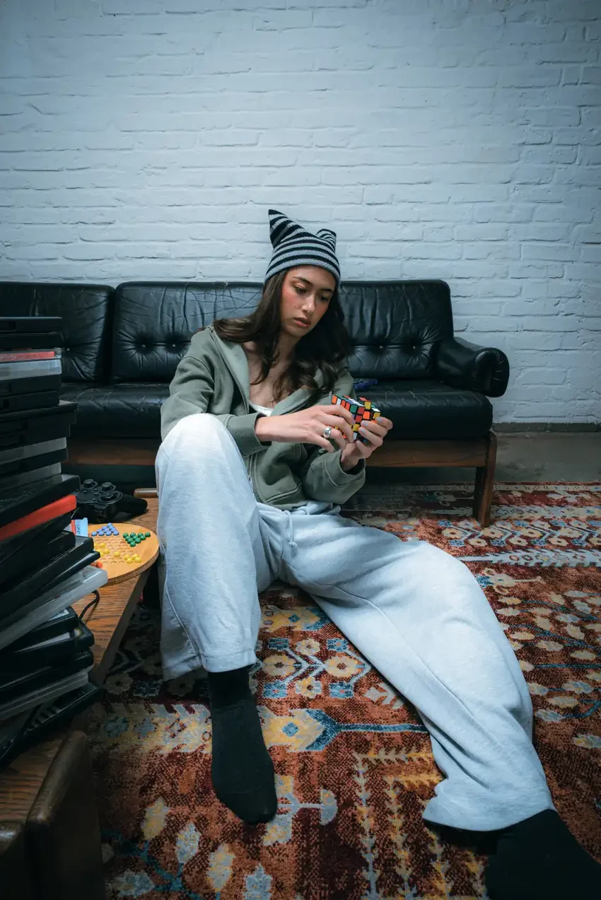 A woman in a striped beanie, olive hoodie, and gray sweatpants sits on a patterned rug turning a colored puzzle cube in both hands.