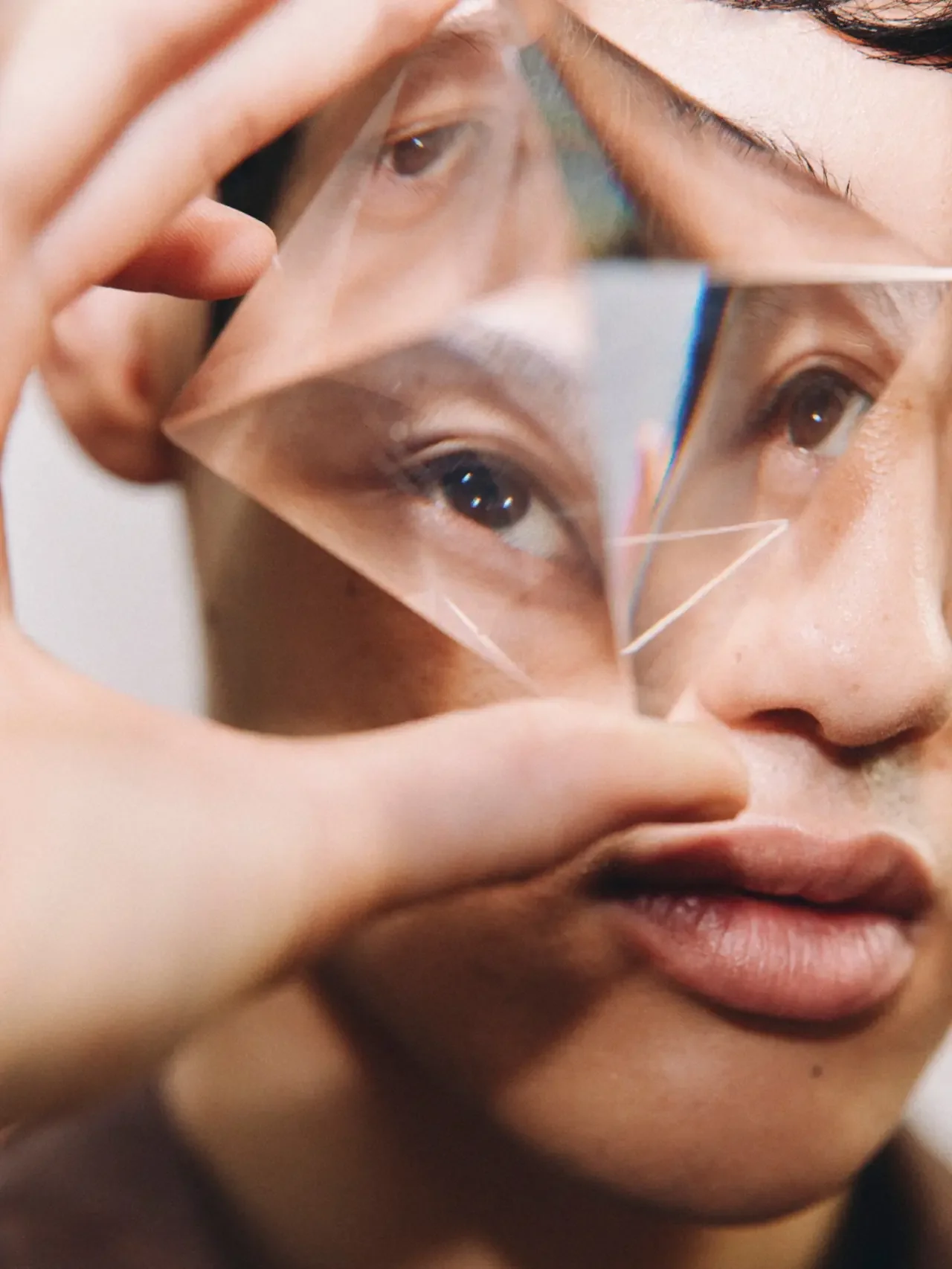 Close-up of a person holding a glass prism in front of their face, with their eyes and nose reflected and split across the angled surfaces.