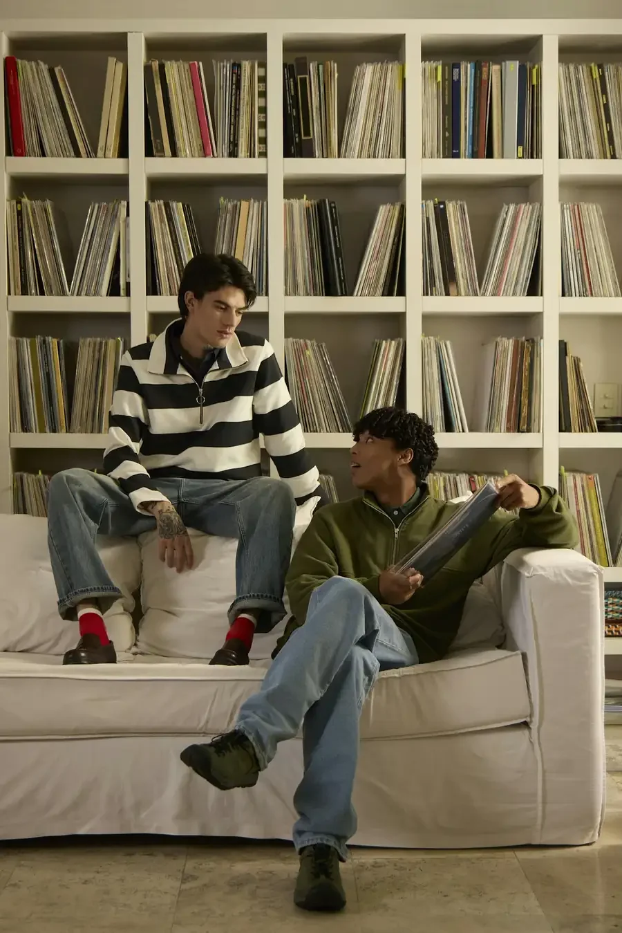 Two friends talking on a couch while one holds a vinyl record, surrounded by full record shelves