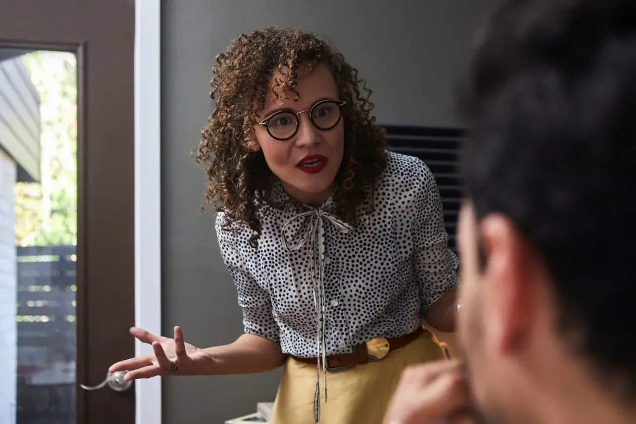 Woman in glasses and a polka-dot blouse leans toward her partner mid-argument, gesturing with frustration as he listens in the foreground.