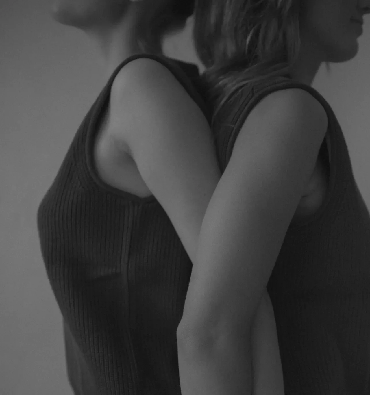 Black-and-white photo of two women in ribbed tank tops standing back to back with their arms interlinked behind them.