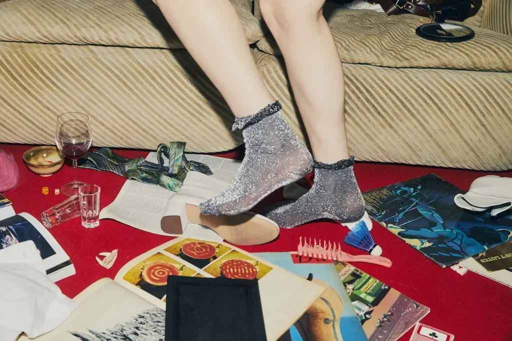 A person's legs in sparkly socks dangle from a couch above a red carpet strewn with open magazines, records, glasses, and scattered objects.