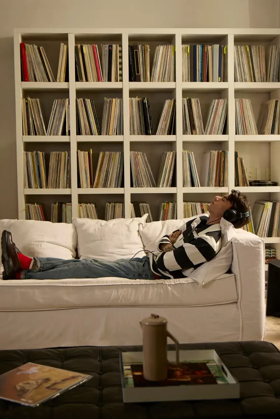 Man lying on a white couch with headphones on, listening to music in front of floor-to-ceiling shelves of vinyl records