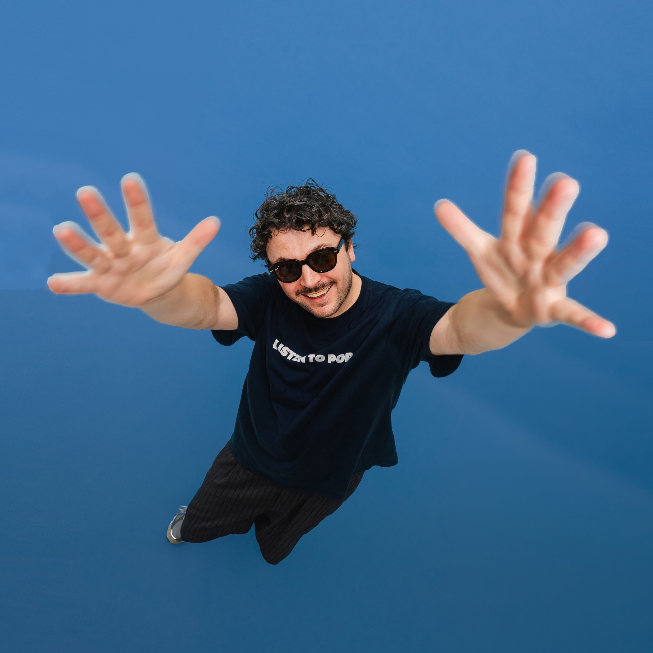 A man with curly hair wearing sunglasses, a black T-shirt, and black pants, smiling and reaching towards the camera against a clear blue sky.