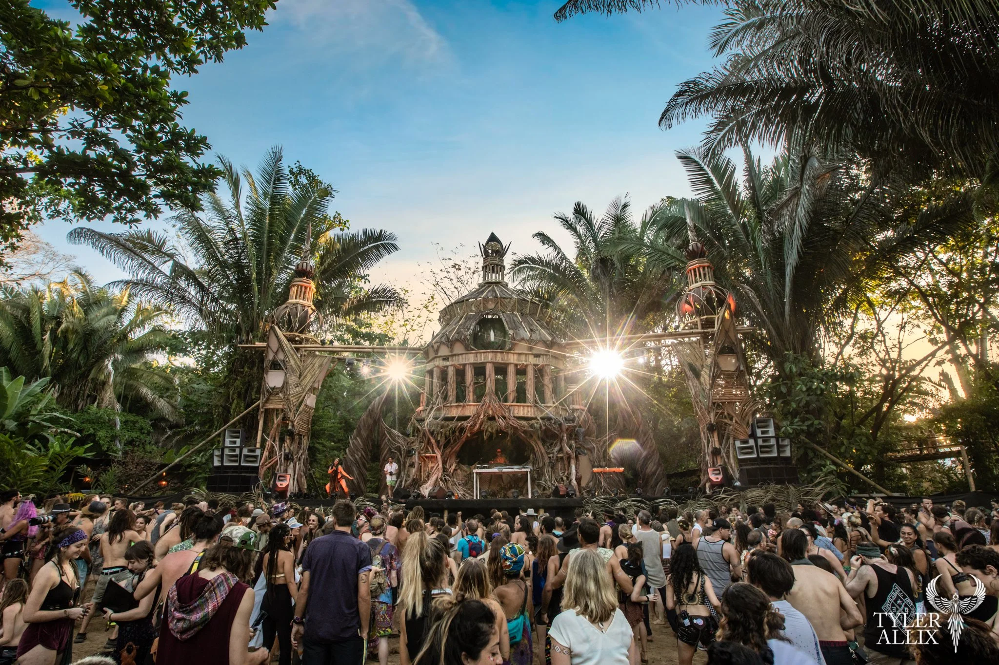 A large outdoor music festival with a wooden stage set amidst lush green trees and palm trees in costa rica, with a crowd of people enjoying the concert during sunset.