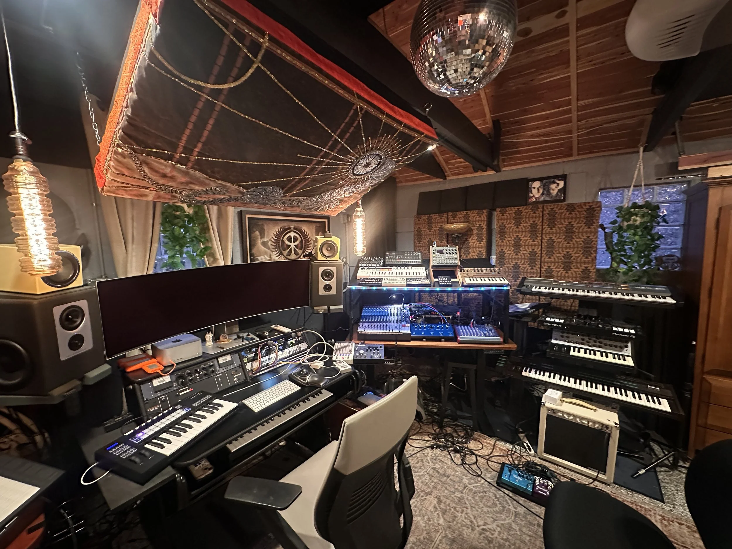 A music studio with various electronic instruments and equipment on desks, including keyboards, synthesizers, and audio mixers, with decorative lighting and wall art in the background.