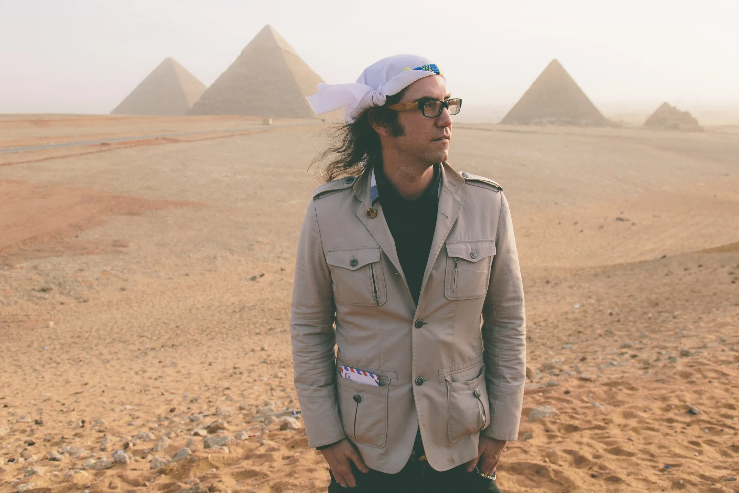 A man with long hair, wearing glasses, a beige jacket, and a white headscarf, standing in a desert with pyramids in the background. Random Rab